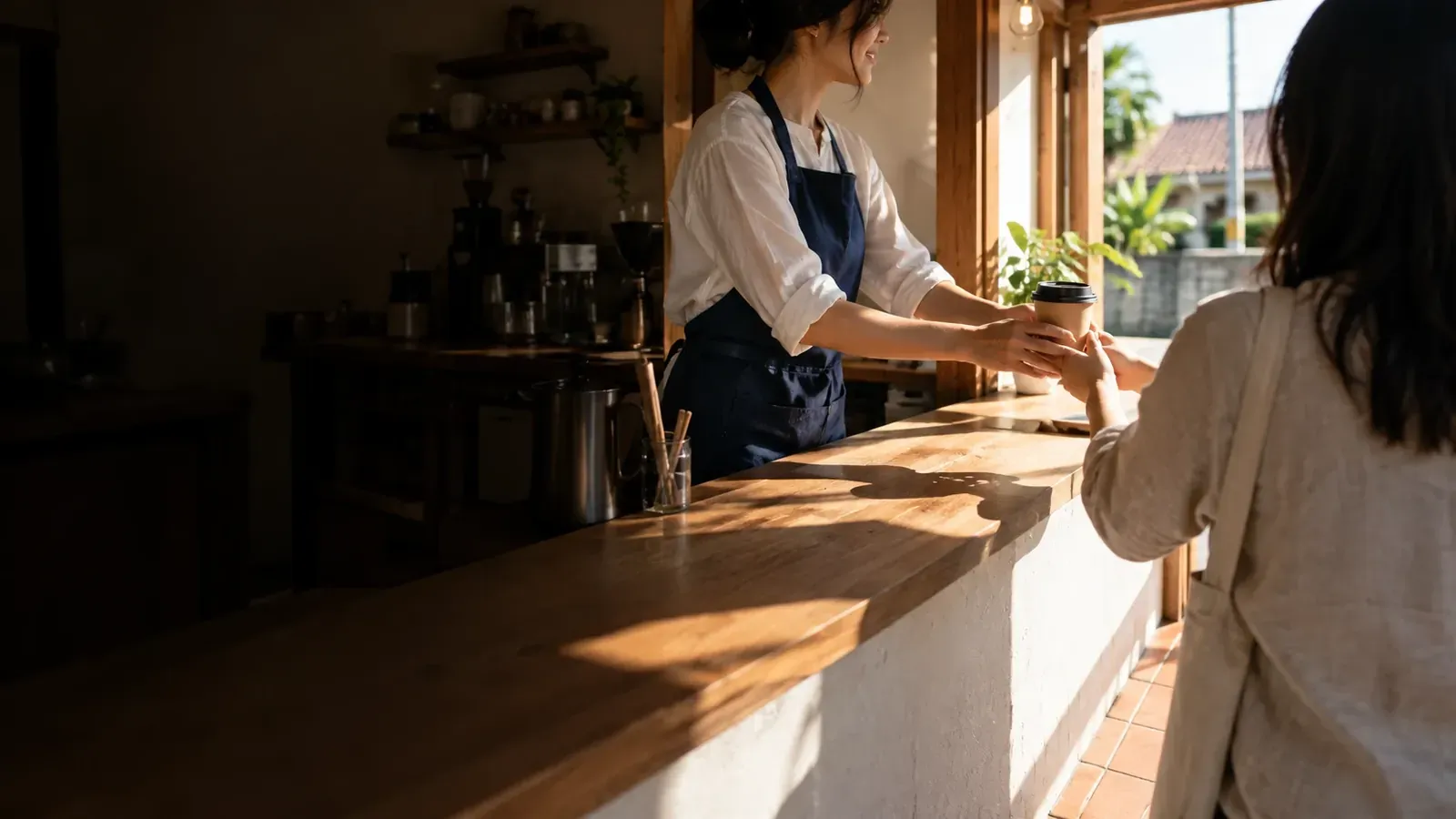 エプロン姿の店員が、カウンター越しにお客さんへテイクアウトのコーヒーを手渡している(AI で生成したイメージ)